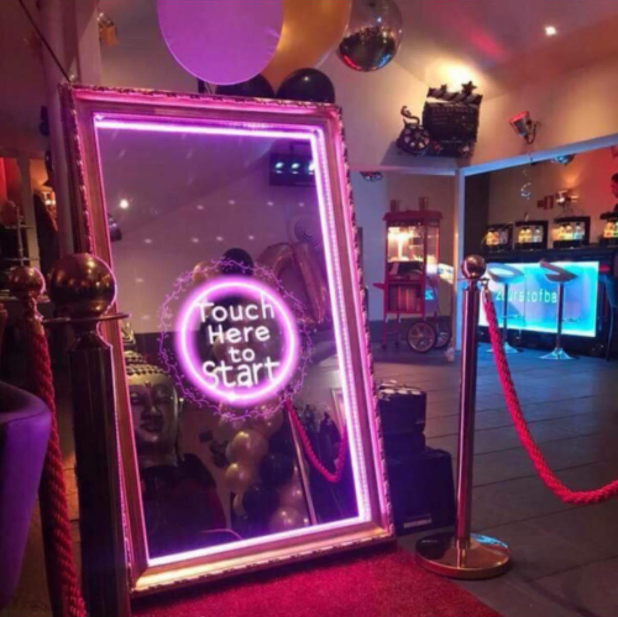 Modern photo booth kiosk with a touchscreen and illuminated ring light, glowing blue and purple in a dimly lit room.