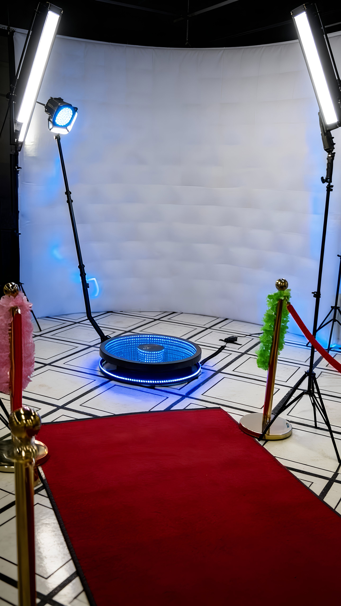 Photo studio setup with red carpet, gold stanchions with colorful feather decorations, softbox lights, and a circular blue LED infinity mirror platform.