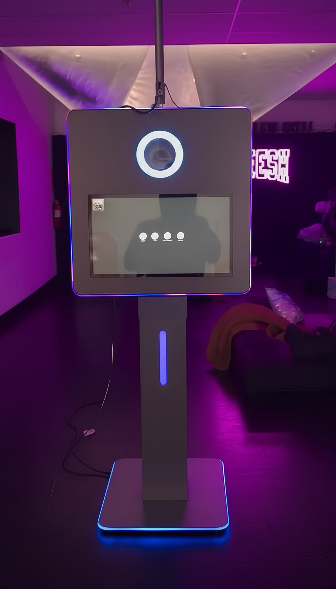 Modern photo booth kiosk with a touchscreen and illuminated ring light, glowing blue and purple in a dimly lit room.