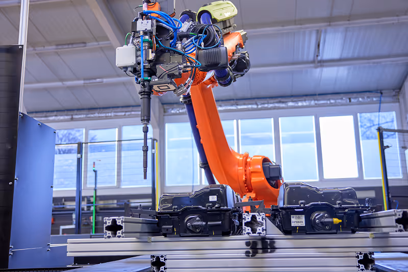 Orange robotic arm equipped with tools working on black mechanical parts on a metal assembly line indoors.