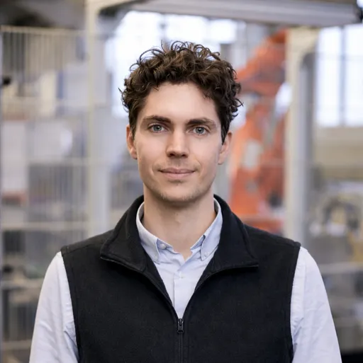 Young man with curly hair wearing a white shirt and black zip-up vest in an industrial setting.