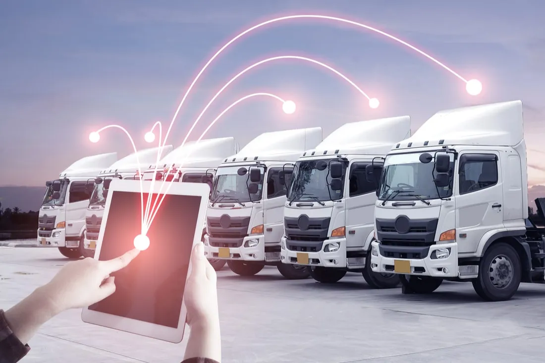 Person using a tablet with digital connections linking to a fleet of white trucks parked in a row at dusk.