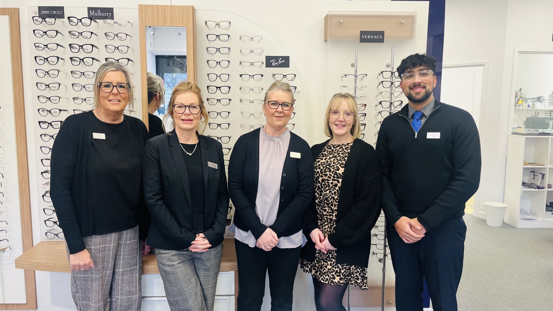 Five optical shop staff members posing together in front of eyeglasses display wall