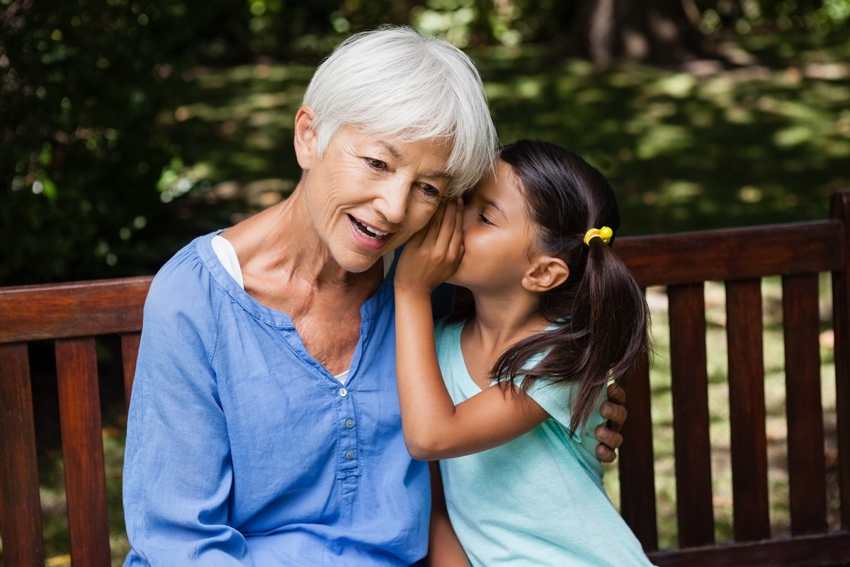 Smiling senior woman with white hair hugs young girl on wooden park bench