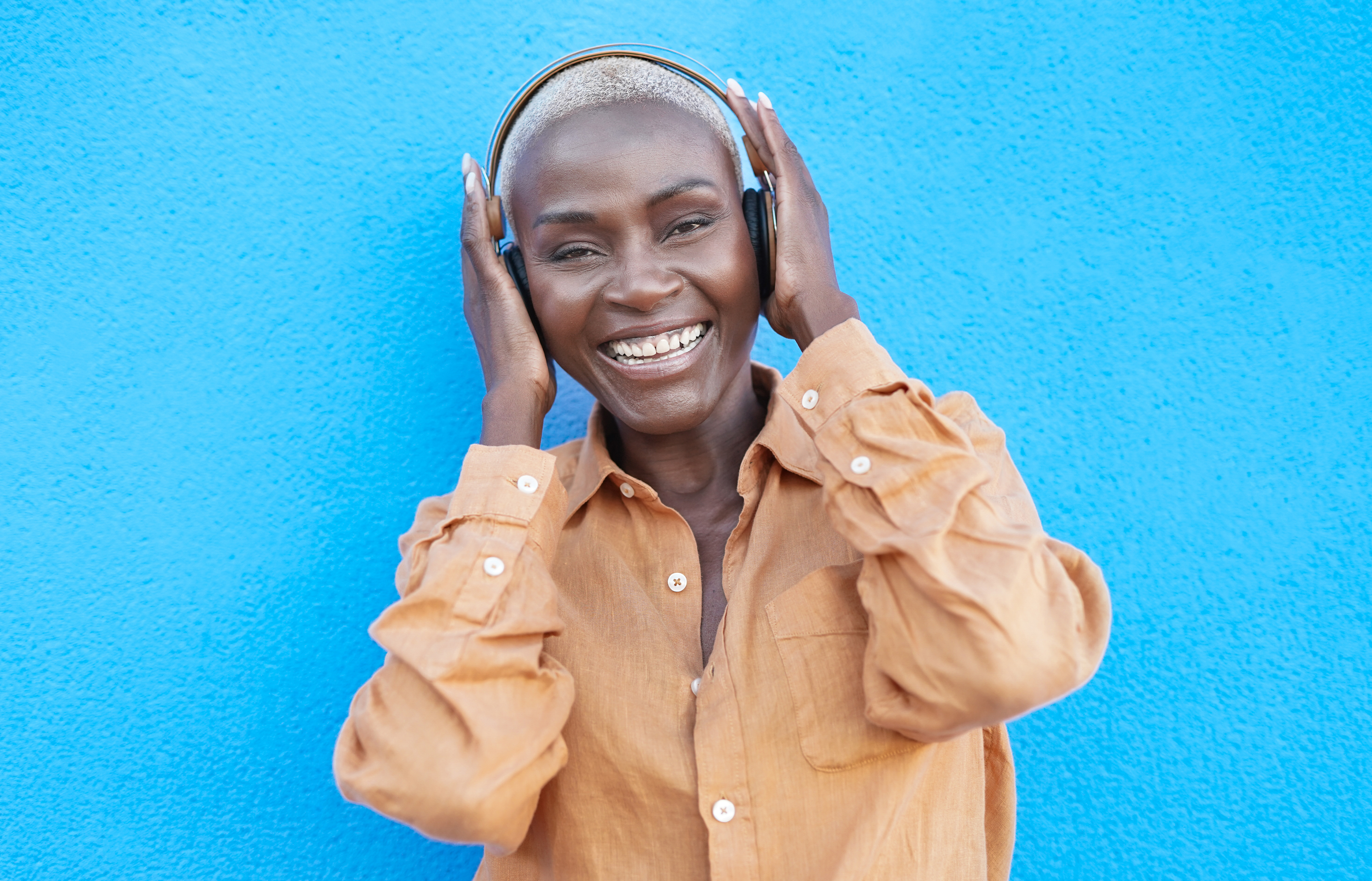 Woman wearing headphones smiling against blue background, wearing tan shirt