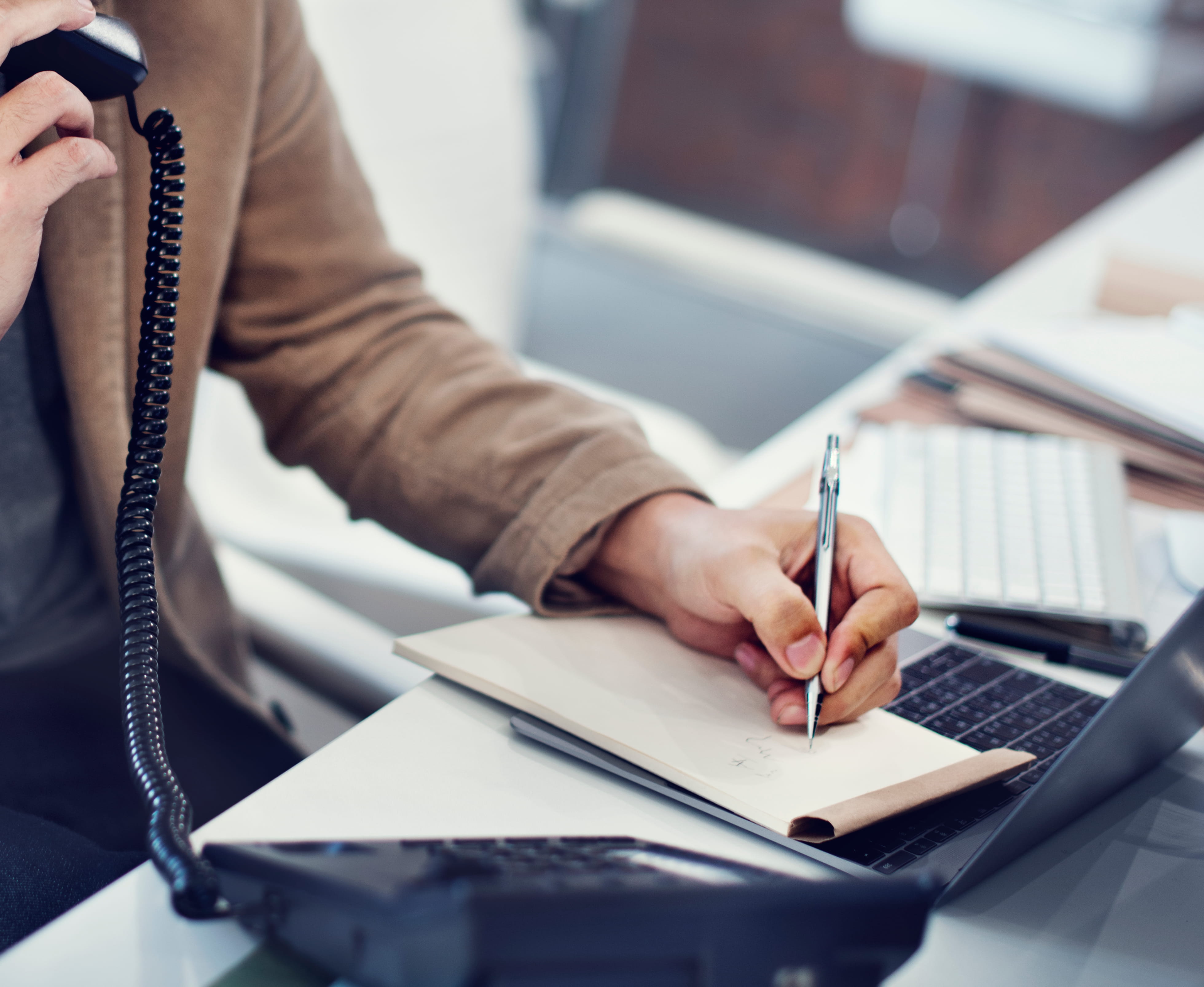 Professional woman writing in notebook while holding telephone handset at desk.