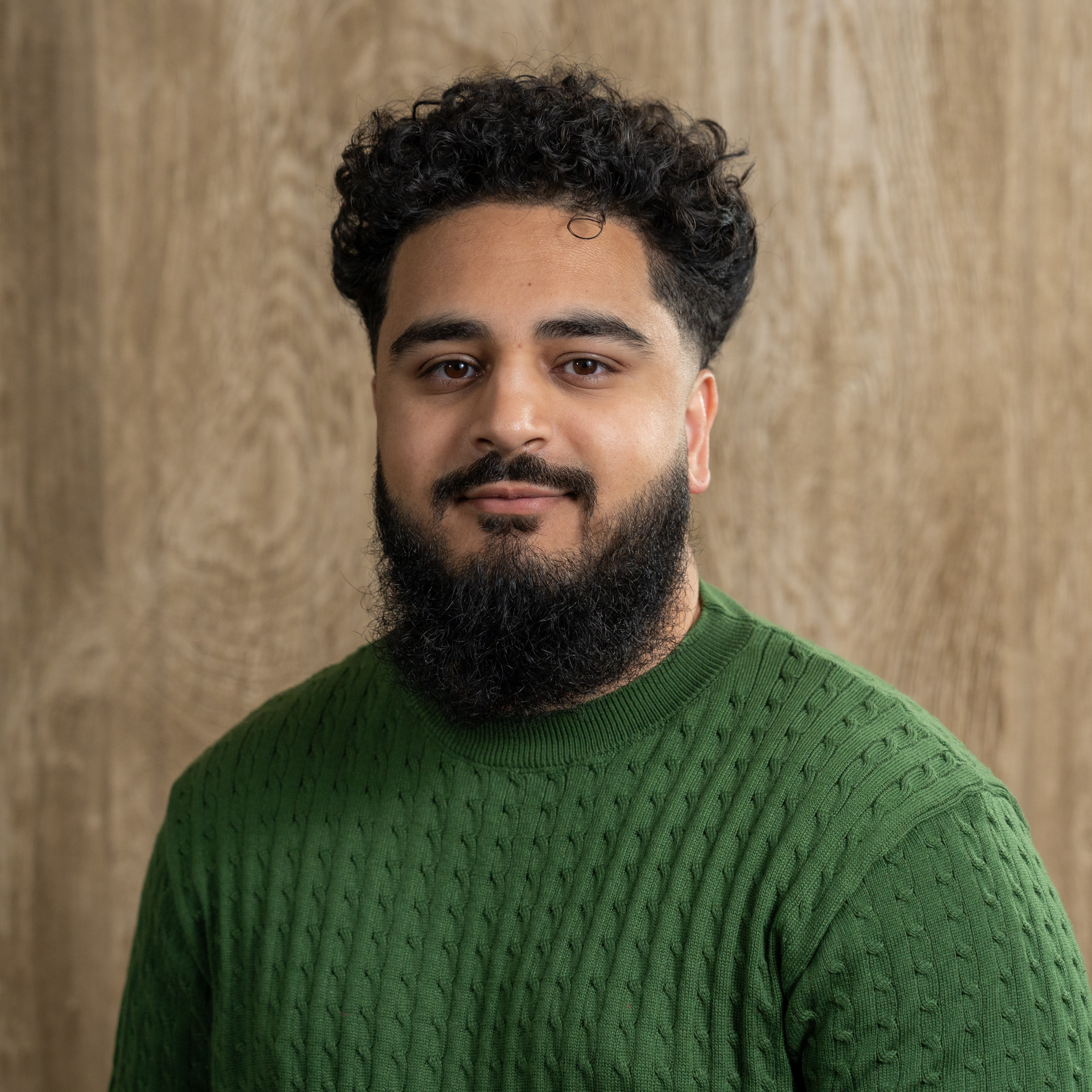 Man with dark curly hair and beard wearing green textured sweater against beige background