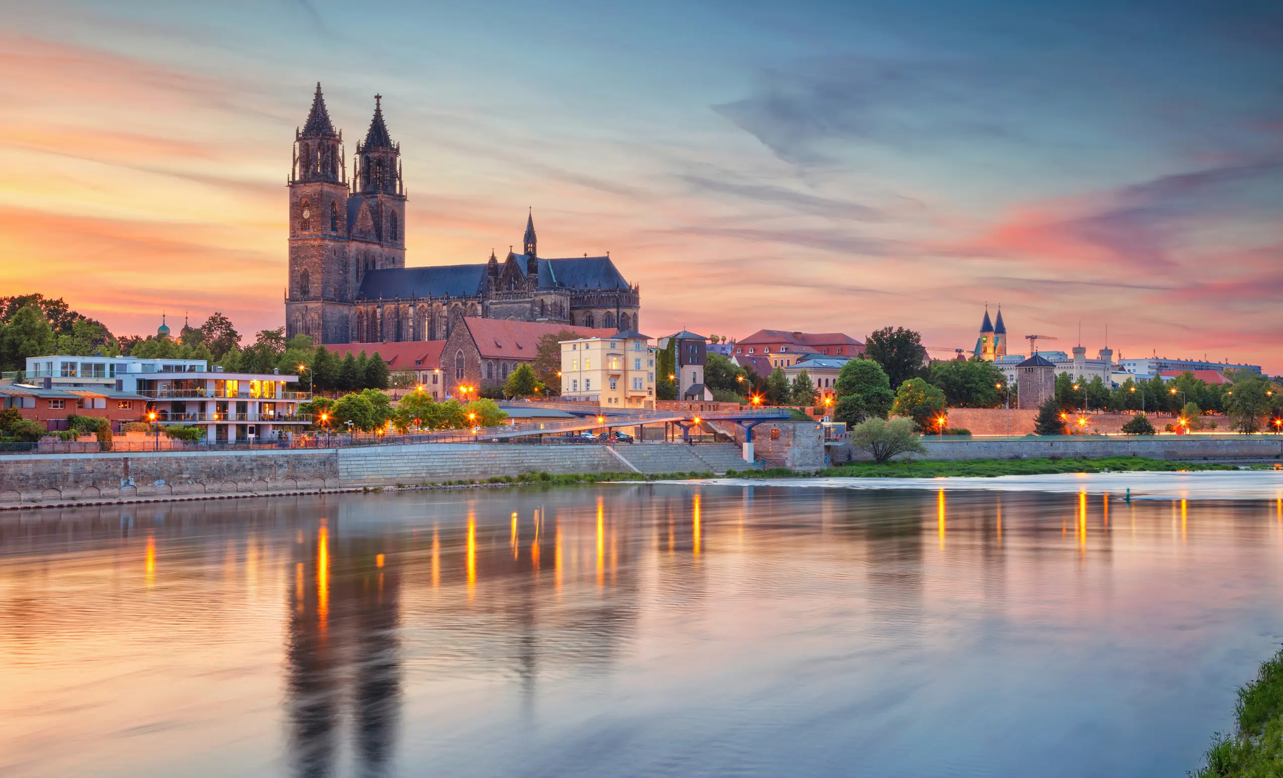 Panoramablick auf die Elbe mit der Silhouette des Magdeburger Doms und umliegenden Gebäuden bei Sonnenuntergang.