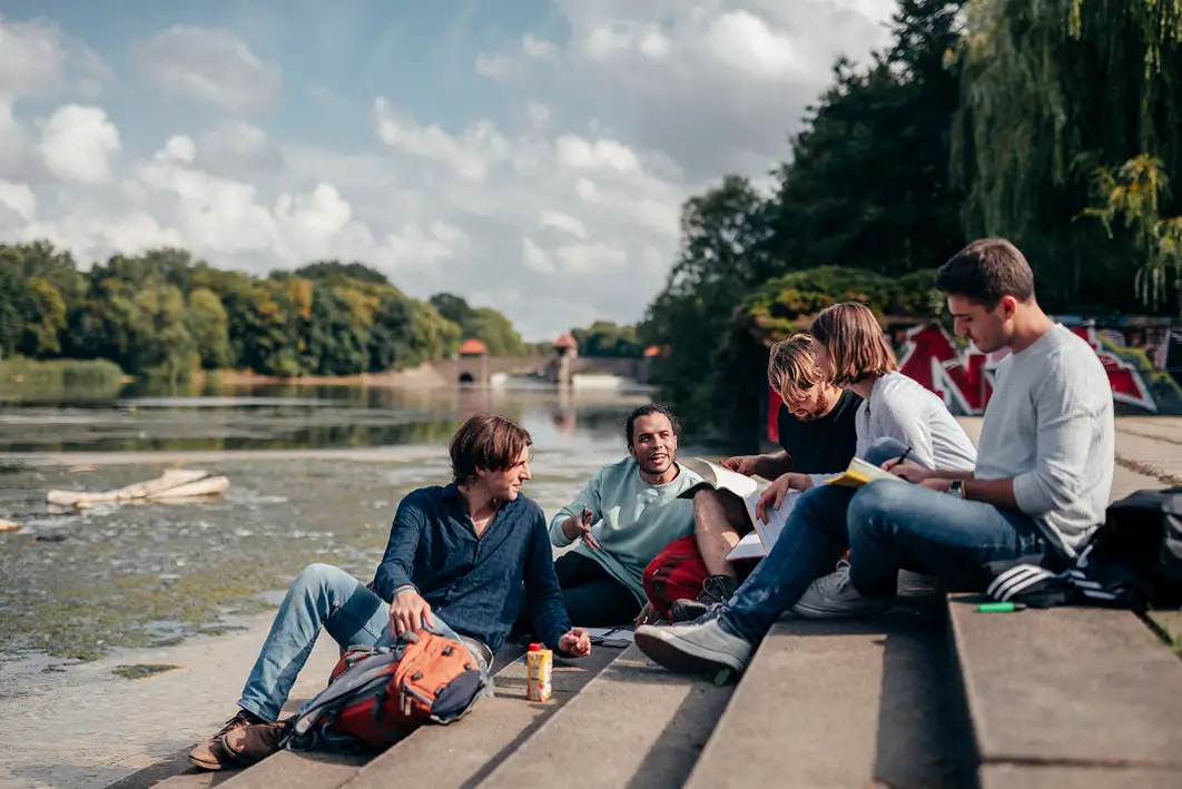 Gruppe von Studierenden sitzt auf Stufen am Ufer eines Flusses mit Rucksäcken und Schreibblöcken und unterhalten sich.