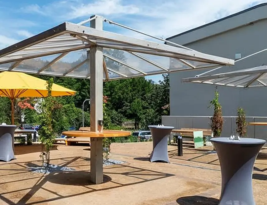 Gepflegter Freiluftbereich mit mehreren Stehtischen, zwei transparenten Sonnenschutzdächern und einem gelben Sonnenschirm.