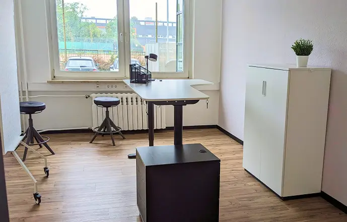 Heller Raum im Beratungscenter mit höhenverstellbarem Tisch, zwei Hockern, einem weißen Schrank mit Pflanze und großen Fenstern mit Blick auf geparkte Autos.