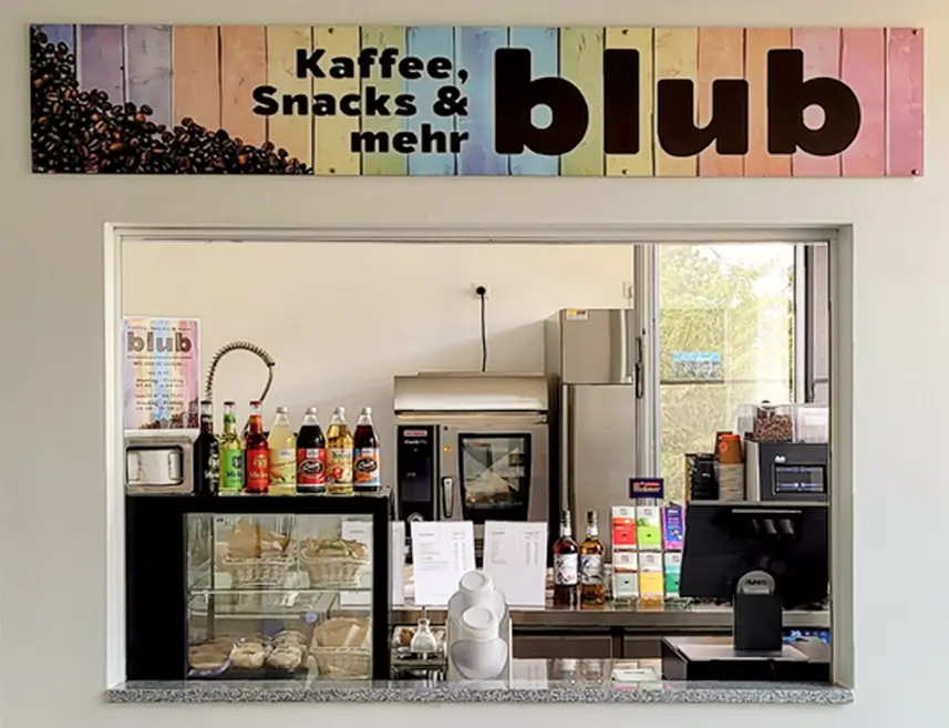 Verkaufstheke mit Kaffeemaschine, Getränken, Snacks und Backwaren unter einem Schild mit der Aufschrift 'Kaffee, Snacks & mehr blub'.