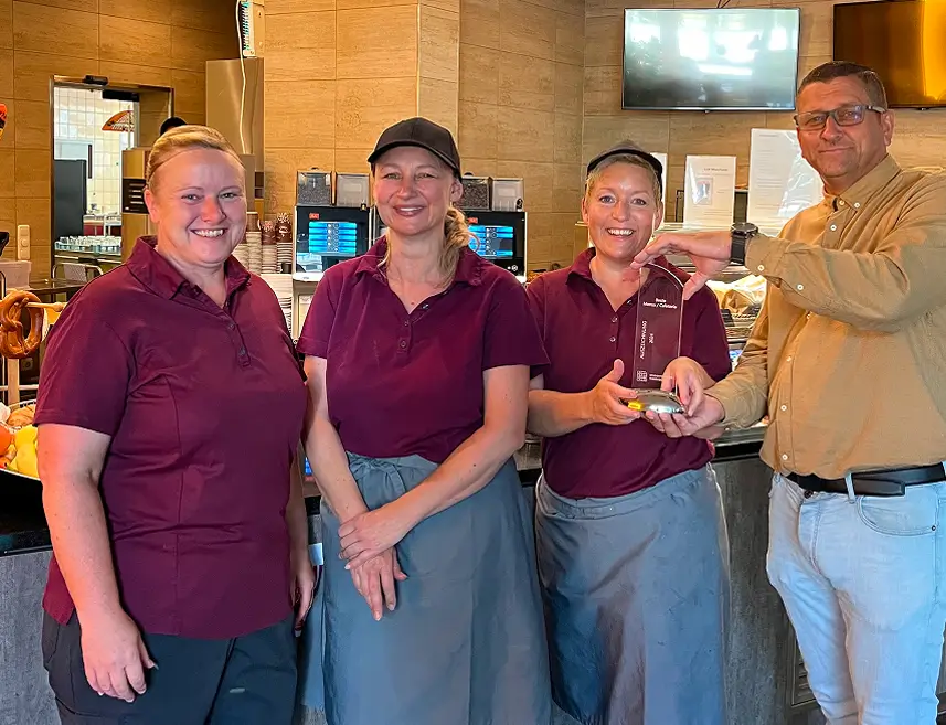 Drei Frauen in einheitlichen dunkelroten Poloshirts und grauen Schürzen stehen nebeneinander in der Mensa und bekommen von einem Mann einen Pokal überreicht.