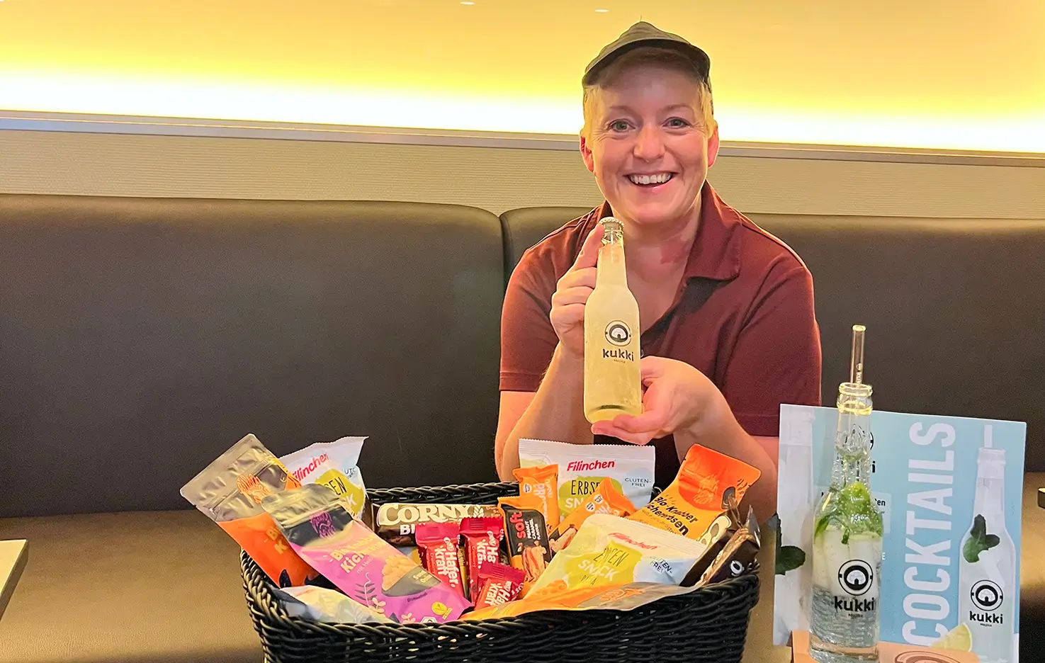 Eine Frau hält eine Flasche Kukki-Cocktail vor einem Korb mit Snacks und einer weiteren Kukki-Cocktailflasche.