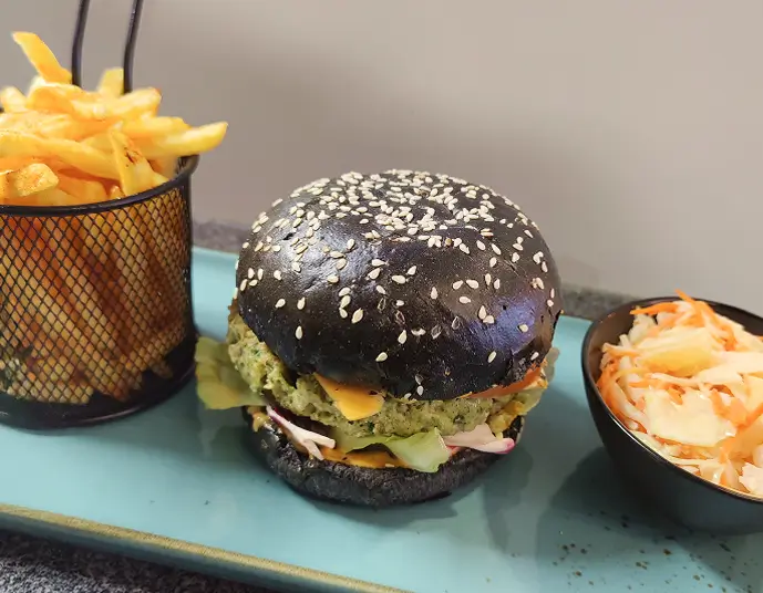 Veggie-Burger mit schwarzem Sesambrötchen, Gemüse-Patty, Käse und Salat, serviert mit Pommes in einem Drahtkorb und Krautsalat in einer schwarzen Schale auf einem Teller.