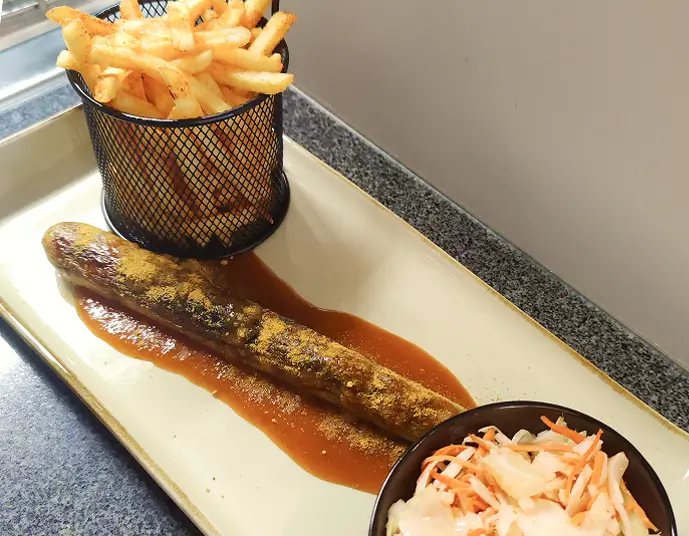 Vegane Currywurst mit Currysoße auf einem Teller, dazu Pommes in einem schwarzen Drahtkorb und Krautsalat in einer kleinen schwarzen Schale.