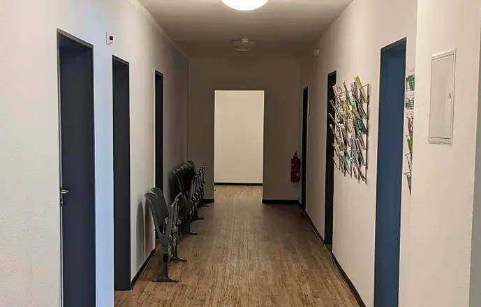 Der Flur im Beratungscenter mit mehreren Türen, links eine Reihe grauer Wartezimmerstühle, rechts eine Wand mit einem befüllten Prospektständer.