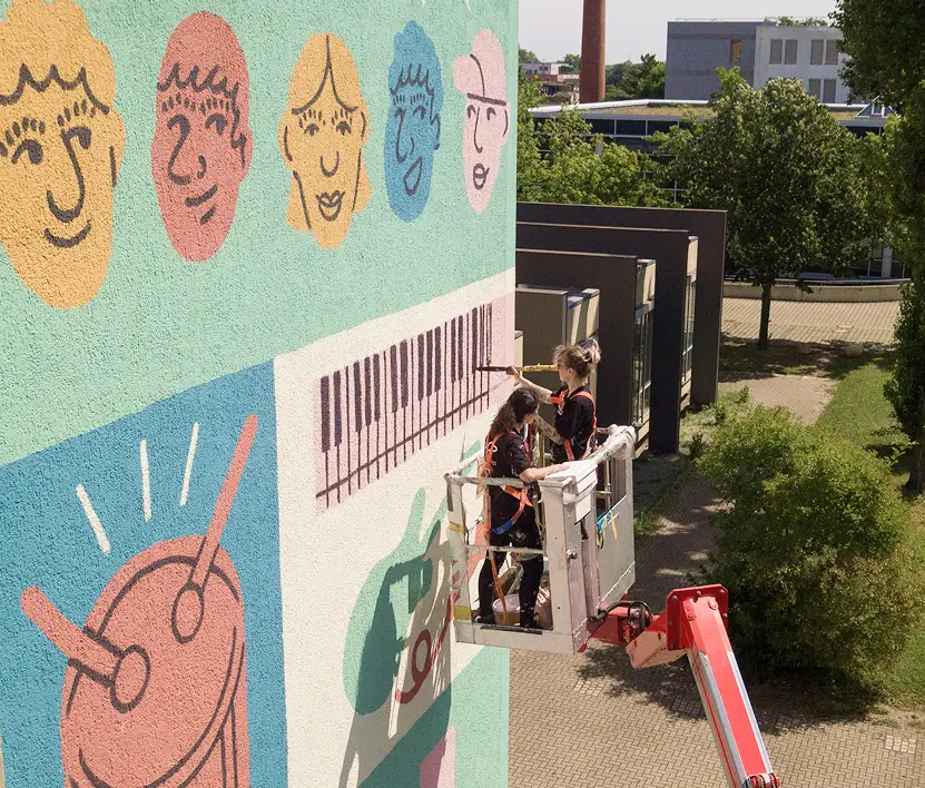 Zwei Personen auf einer Hebebühne gestalten eine bunte Fassade mit stilisierten Gesichtern, einem Klavier und einer Trommel.