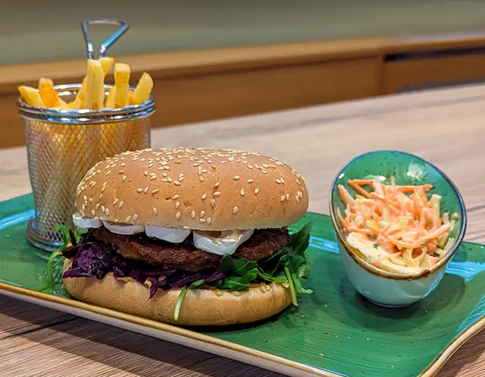 Burger mit Sesambrötchen, Salat, Camembert und Rotkohl, dazu Pommes in einem kleinen Metallkorb und ein Becher mit Krautsalat auf einem grünen Teller.