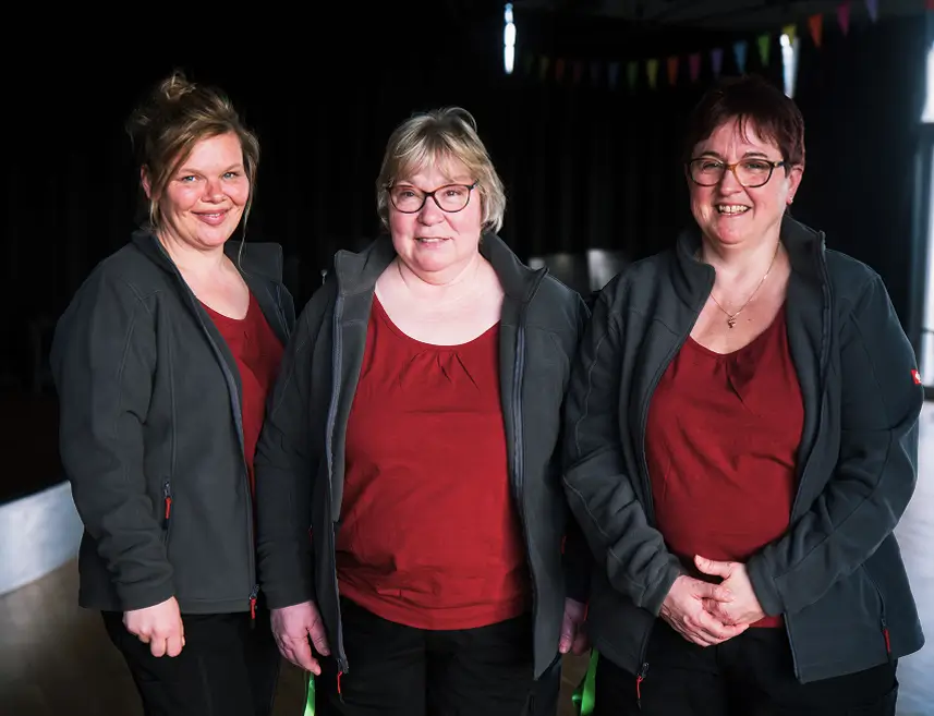 Drei Frauen stehen nebeneinander, lachen in die Kamera und tragen einheitliche Klamotten: ein dunkelrotes T-Shirt, eine graue Weste und eine schwarze Hose.