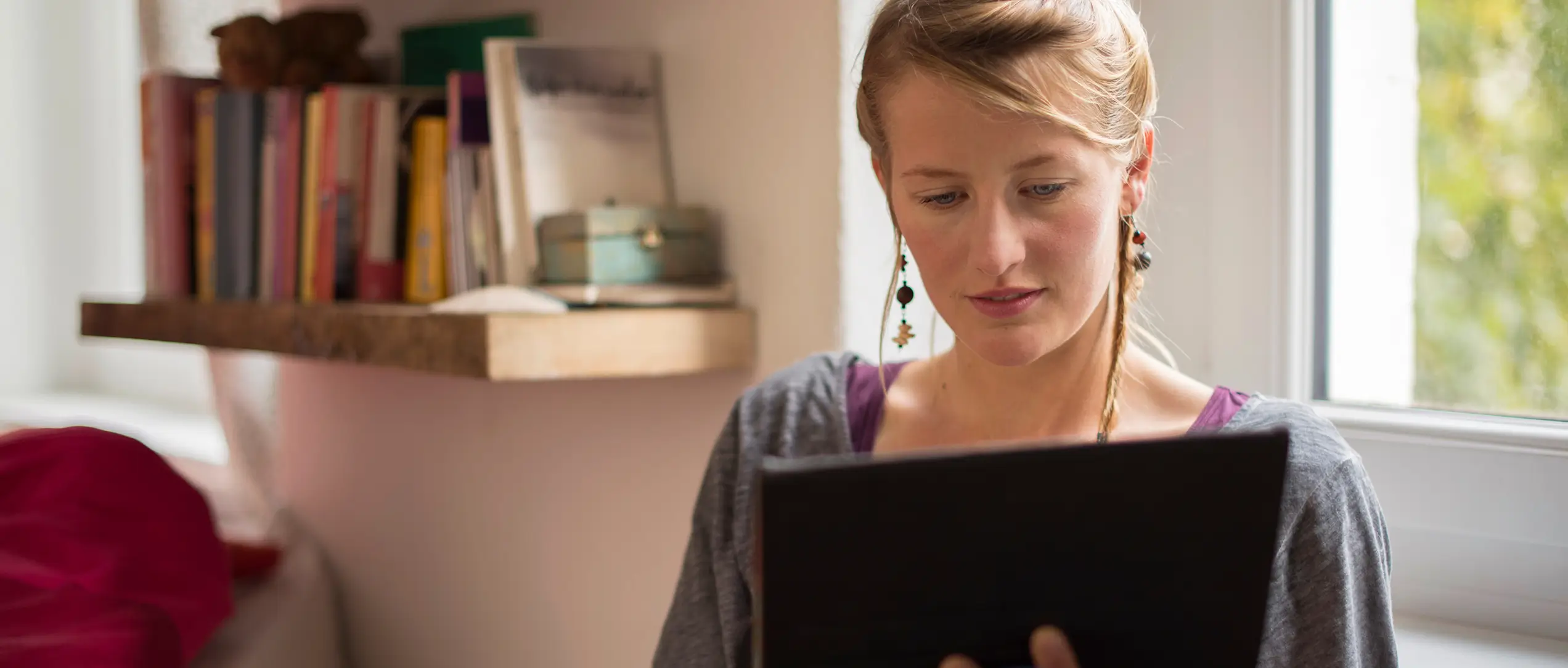Studentin sitzt mit Tablet vor einem Fenster in einem Raum mit Bücherregal.