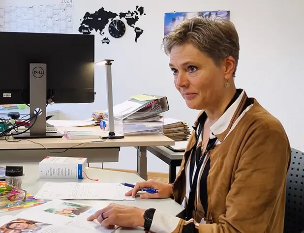 Andrea Köhler steht an einem Schreibtisch, zeigt auf ein aufgeschlagenes Magazin und scheint zu einer weiteren Person hinter der Kamera zu sprechen.