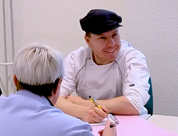 Sebastian Braun sitzt mit weiteren Personen an einem Tisch, er trägt Kochkleidung und eine Kochmütze und hält einen Stift in der Hand.