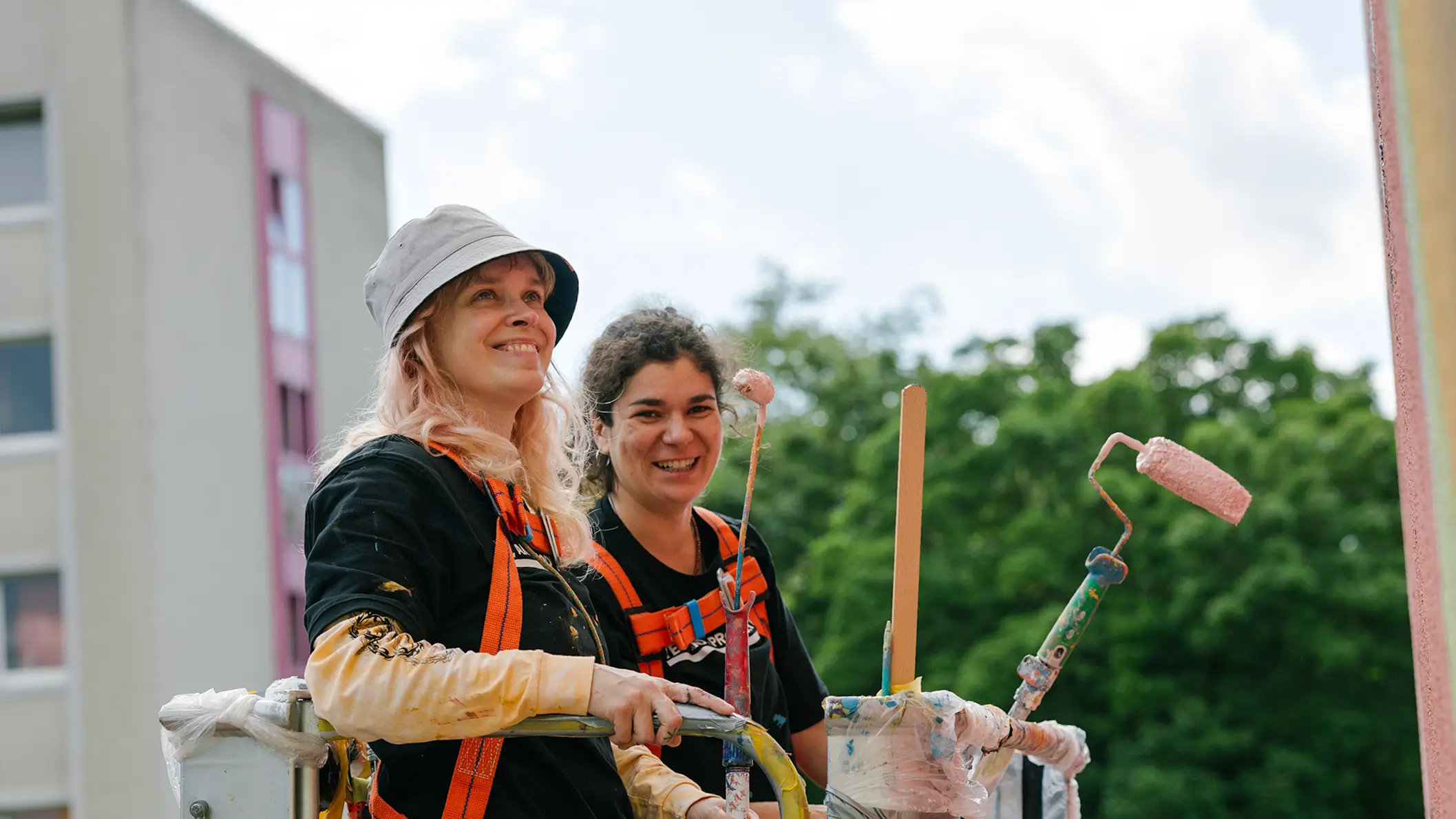 Zwei Personen in Arbeitskleidung mit Farbrollen vor einer Fassade auf einer Hebebühne, im Hintergrund Bäume und ein weiteres Gebäude.