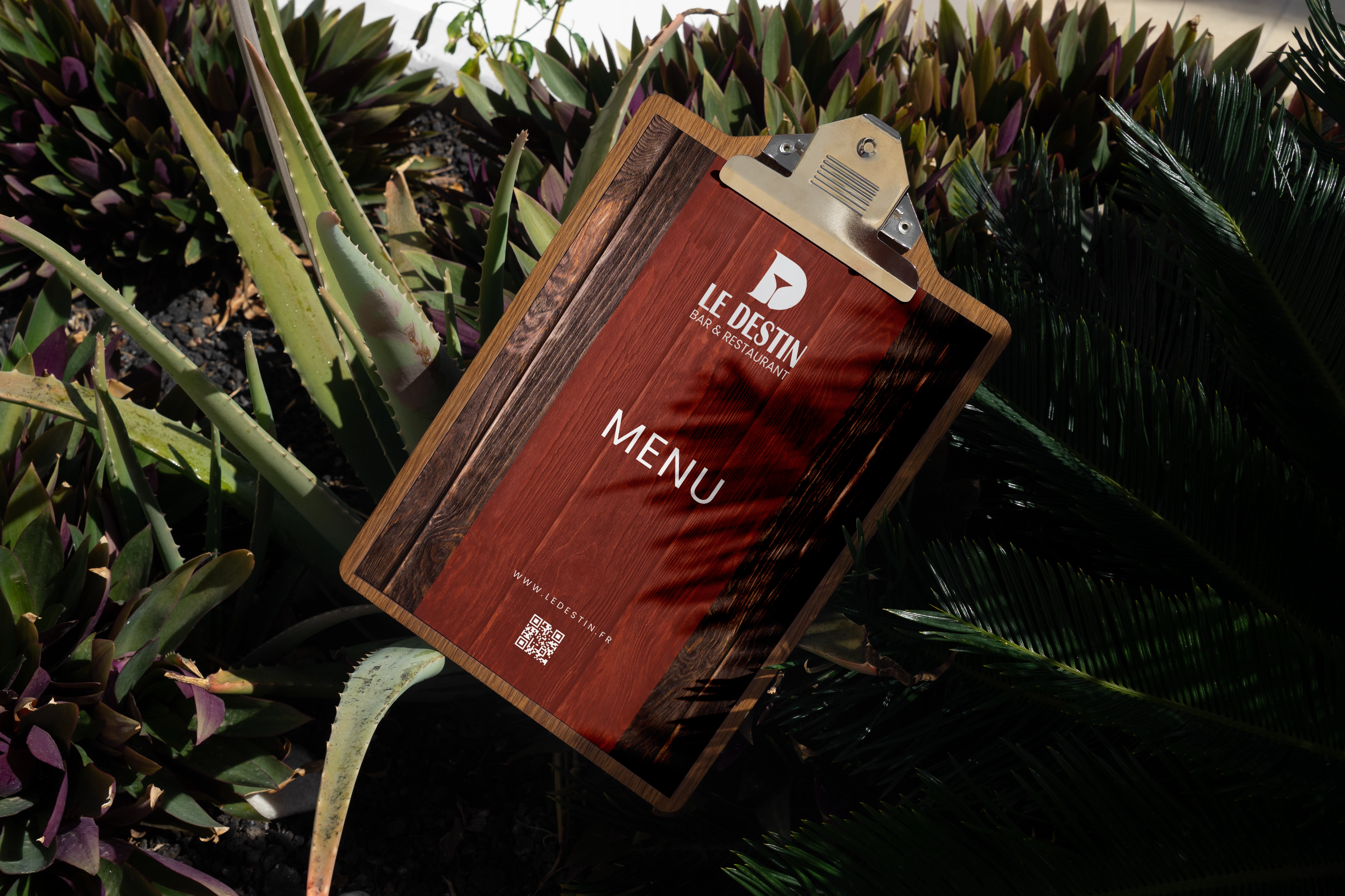 Menu Le Destin Bar & Restaurant sur une planche en bois posée parmi des plantes vertes.