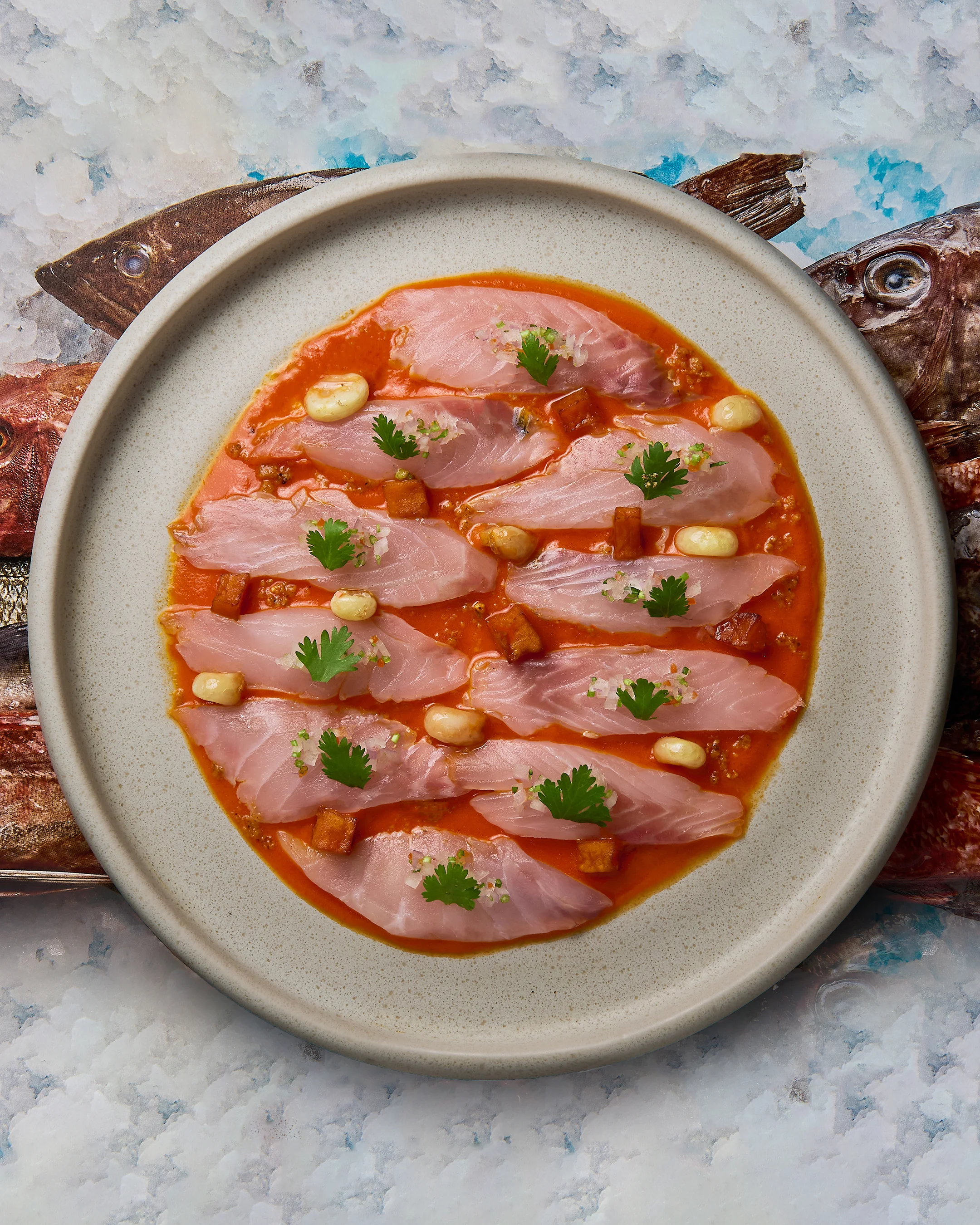 Plate of tiradito with thin fish slices, red rocoto sauce, corn kernels, fried potato cubes, and cilantro leaves.
