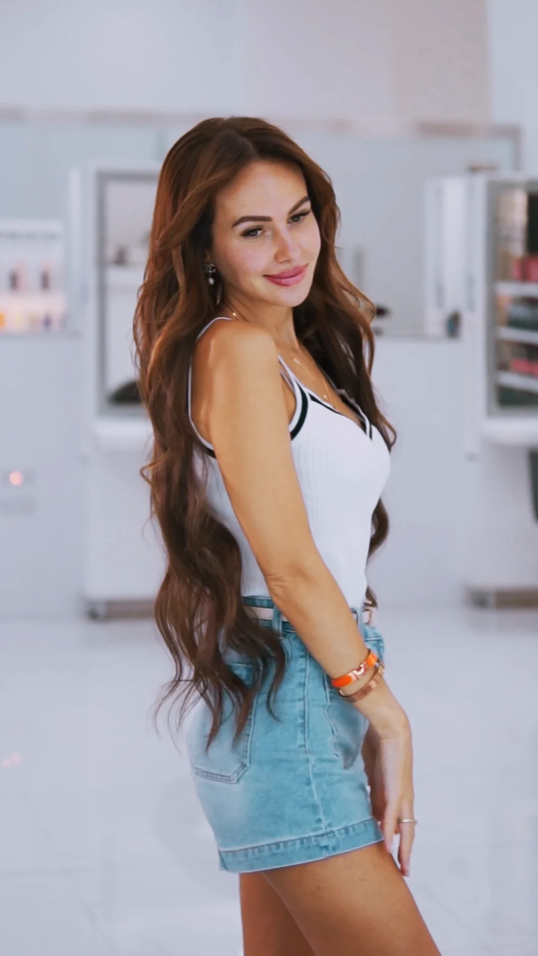 Smiling woman with long wavy brown hair wearing a white tank top and denim skirt standing in a bright indoor space.