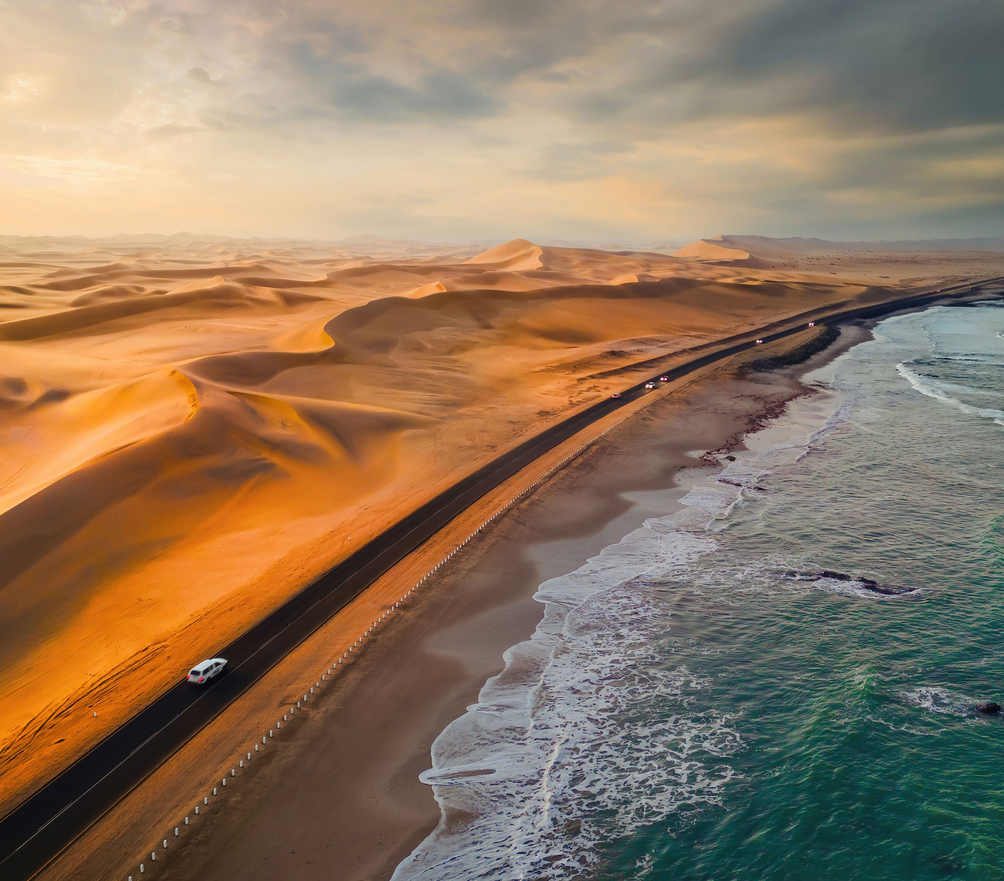 Coastal road running between orange sand dunes and a blue ocean under a cloudy sky at sunset.