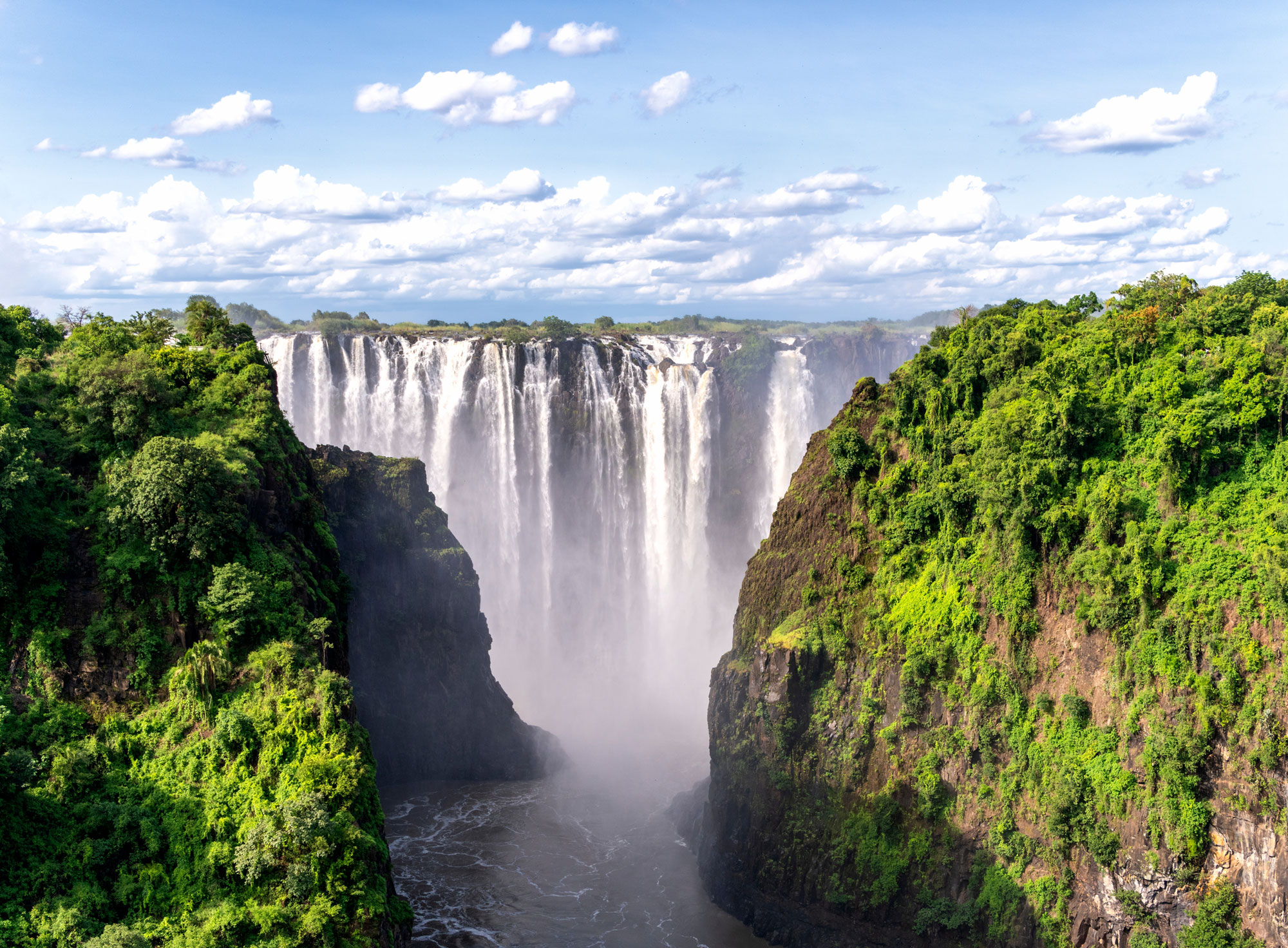 Victoria Falls waterfall cascading between lush green cliffs under a blue sky with scattered clouds.