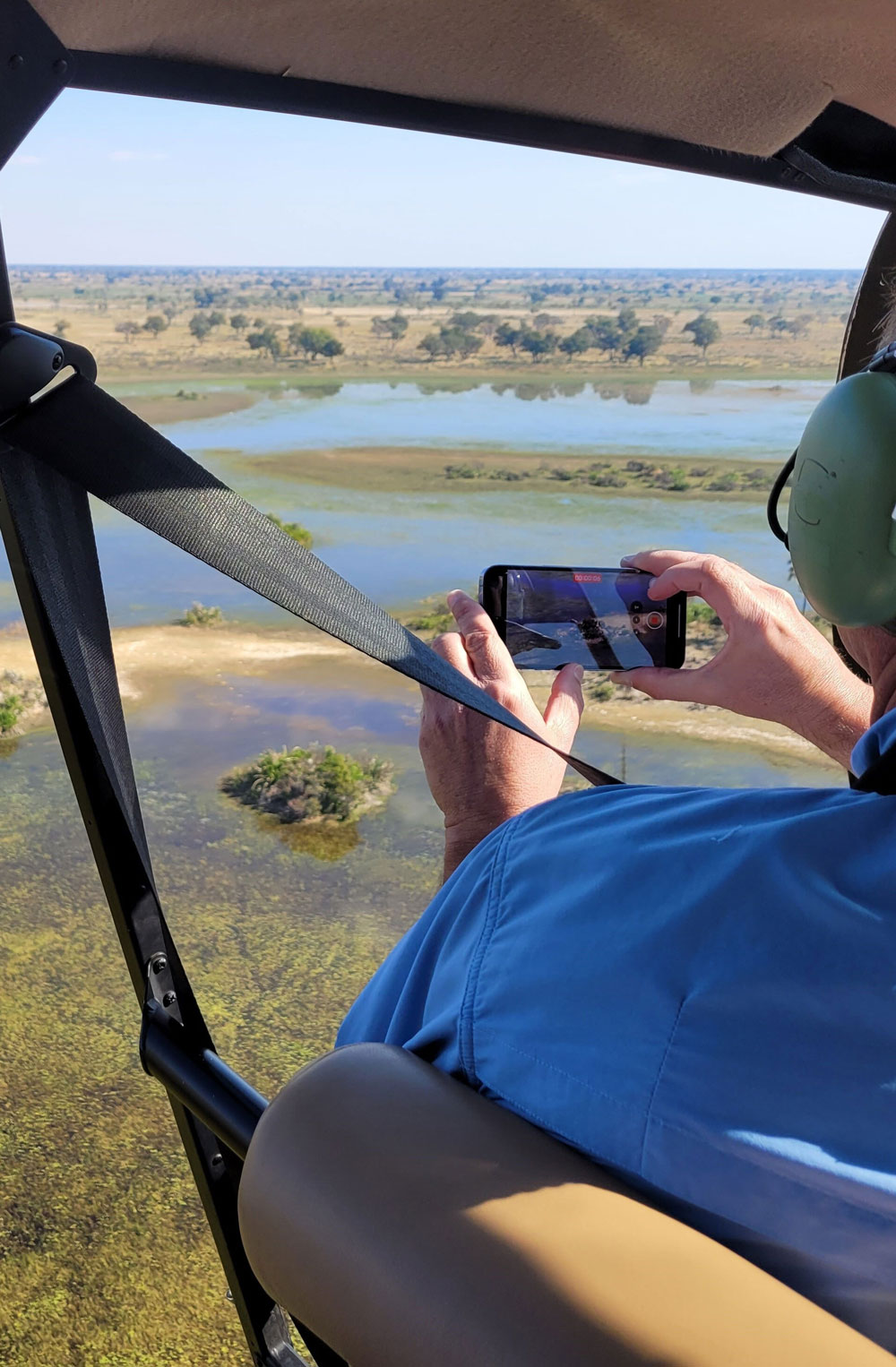 Person wearing headphones and blue shirt filming a wetland landscape from inside a helicopter.