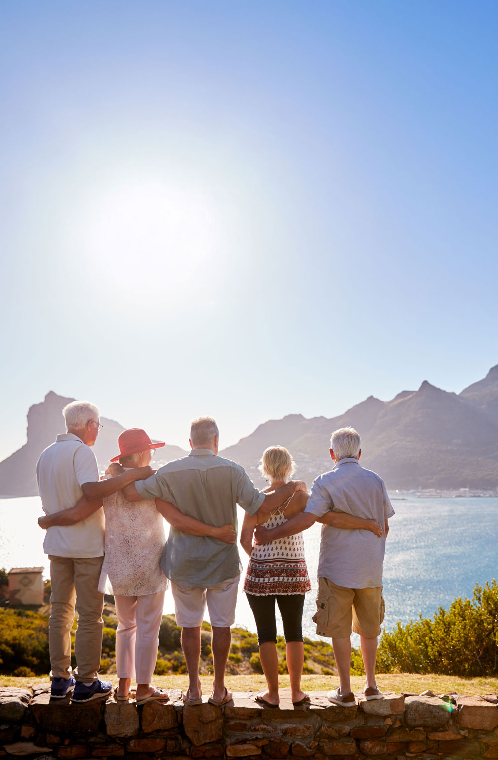 Five older adults standing arm in arm on a stone wall overlooking a sunny coastal landscape with mountains and water.