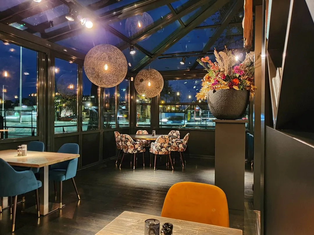 Gezellige restaurantruimte 's nachts met glazen dak, verschillende kleurige stoelen en grote vaas met bloemen.