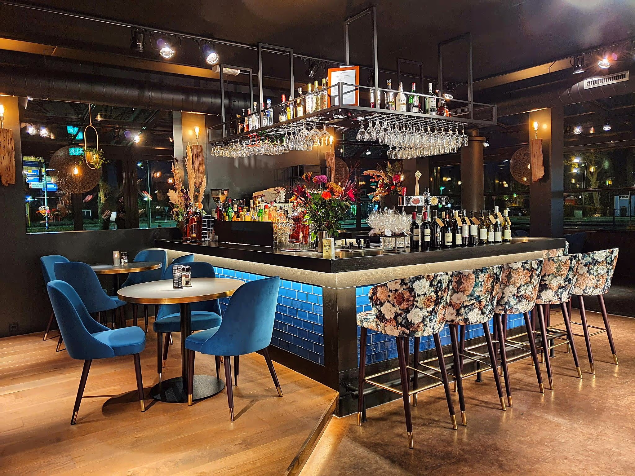 Gezellige bar met blauwe stoelen rond houten tafels en bloemrijke barkrukken bij een bar met blauwe tegels en glazen boven de bar.