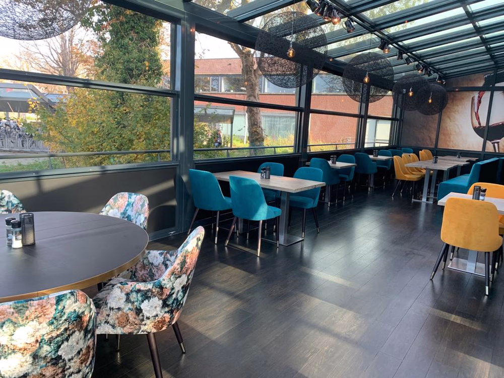Binnenruimte van een modern restaurant met glazen wanden en plafonds, verschillende tafels met blauwe, gele en bloemenpatroon stoelen.