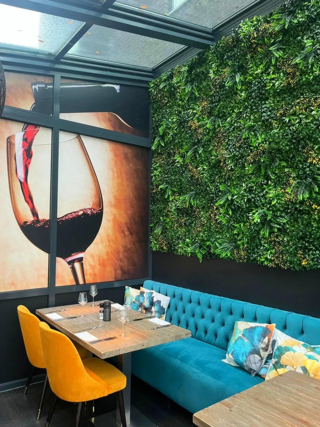 Restaurantinterieur met een houten tafel, twee gele stoelen, een blauwe bank met bloemkussens, een groene plantenmuur en een foto van rode wijn die in een glas wordt geschonken.