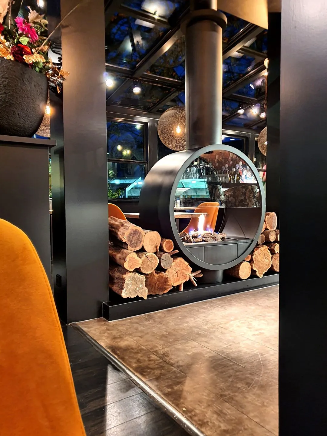 Moderne open haard met brandend vuur en gestapelde houtblokken in een stijlvol restaurant met oranje stoelen.