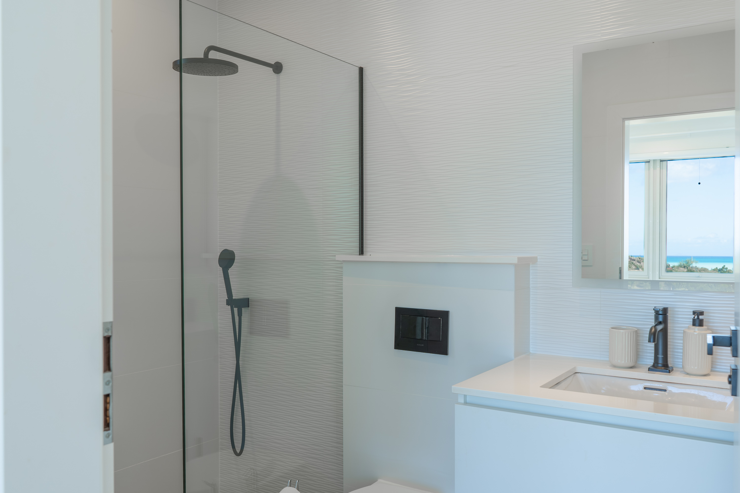 Modern white bathroom with glass shower enclosure, black fixtures, and a vanity with sink and mirror reflecting a window with ocean view.