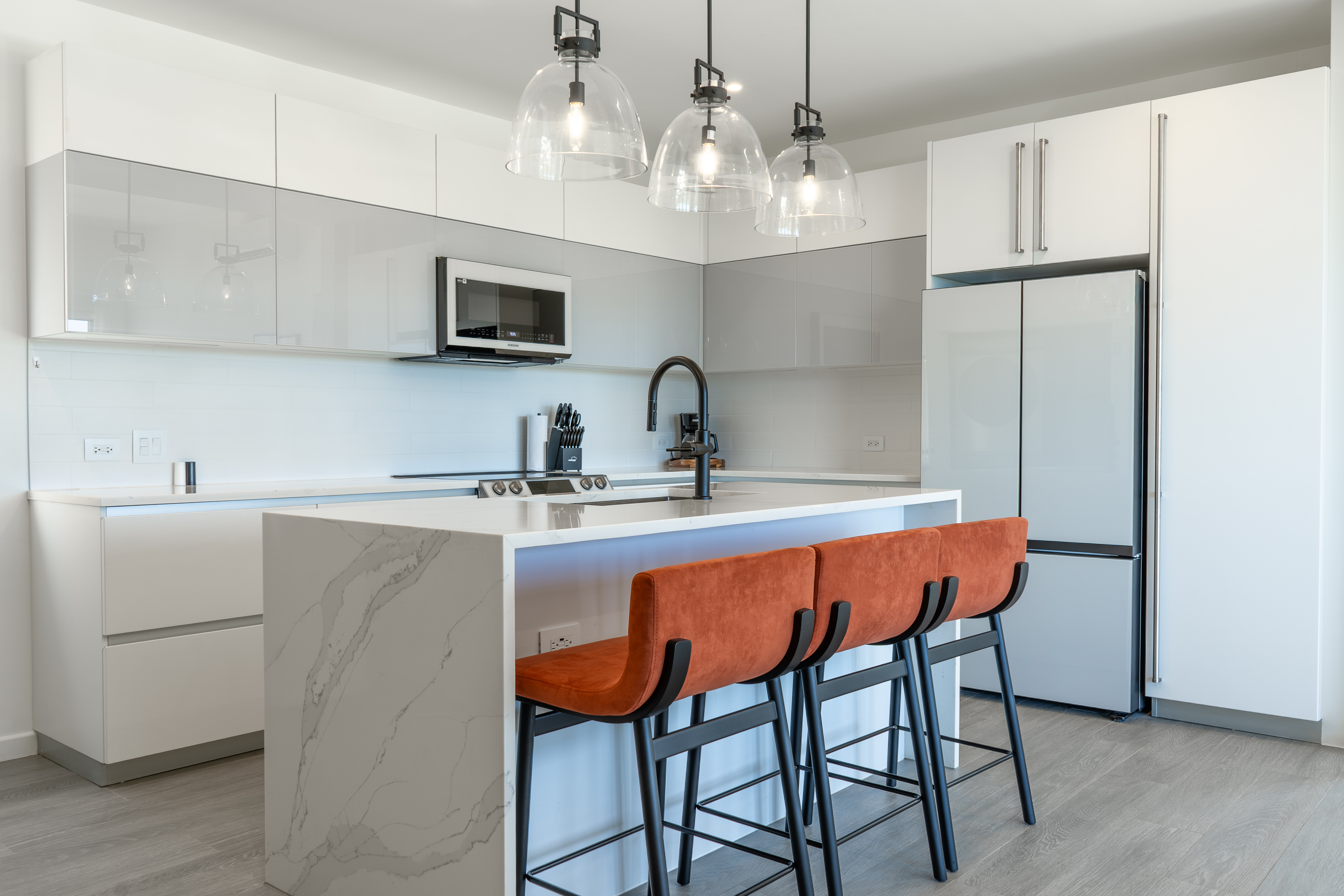 Modern white kitchen with marble island, three orange bar stools, black faucet, and stainless steel appliances.