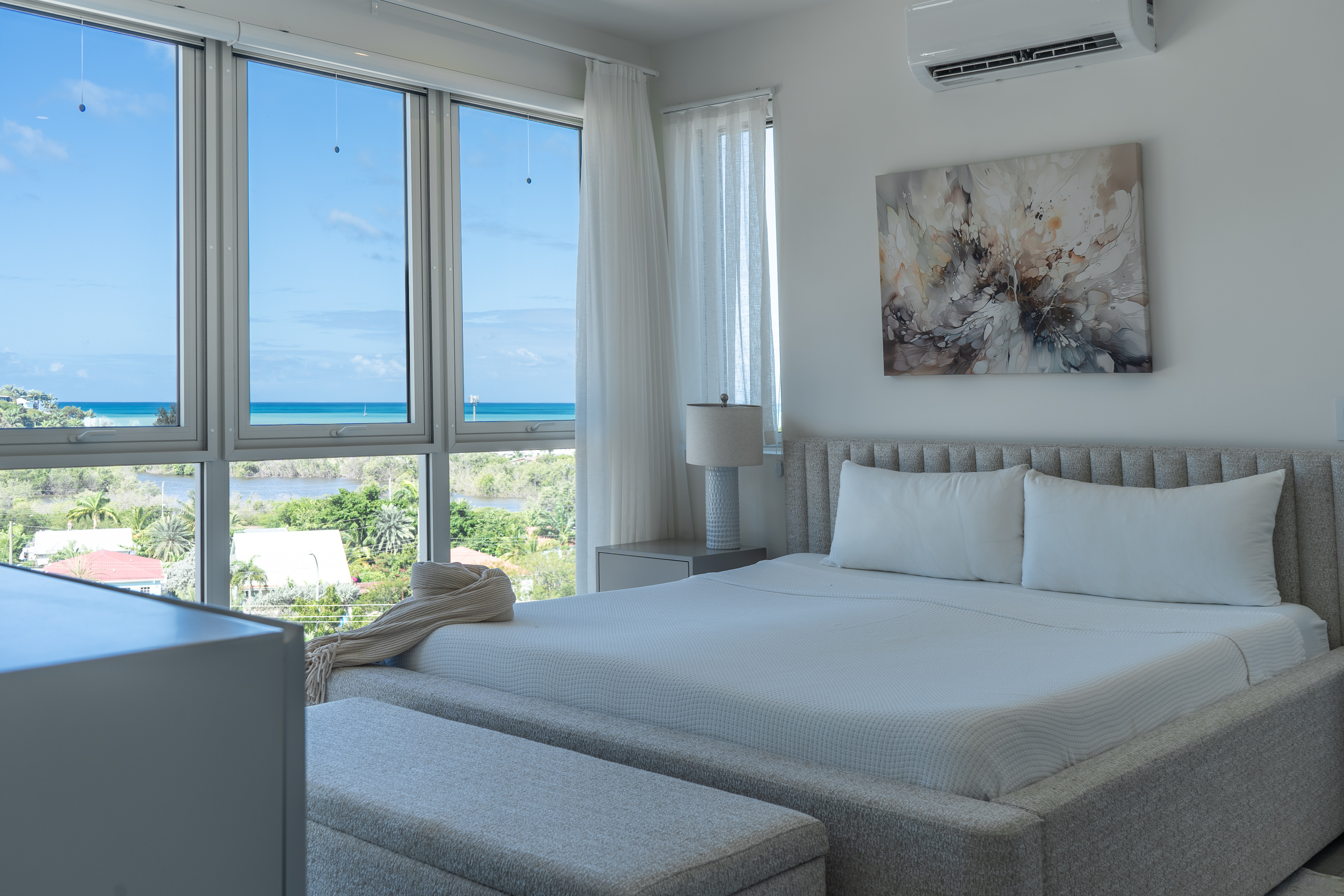 Modern bedroom with large windows overlooking a coastal landscape, featuring a gray upholstered bed with white bedding and pillows, a bedside lamp, and a decorative abstract painting on the wall.