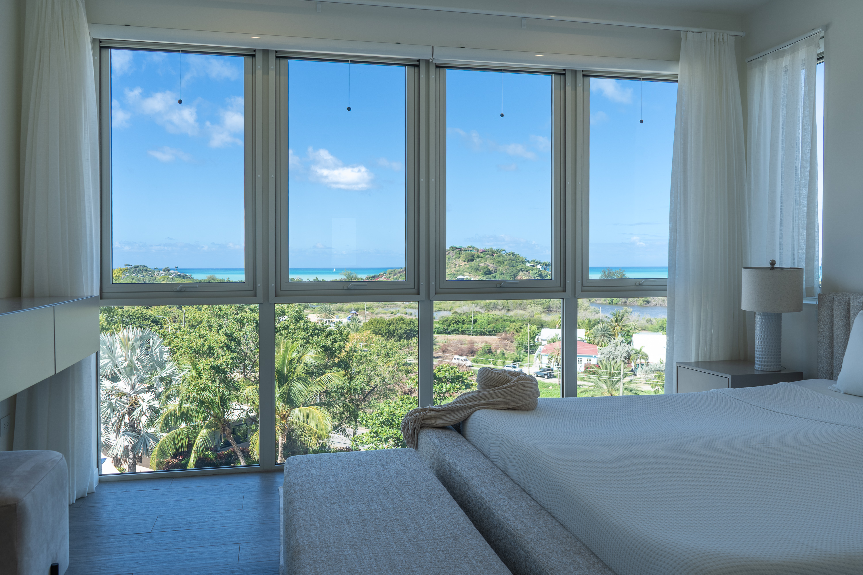 Bright bedroom with large windows showing a view of green trees, hills, and blue ocean under a clear sky.