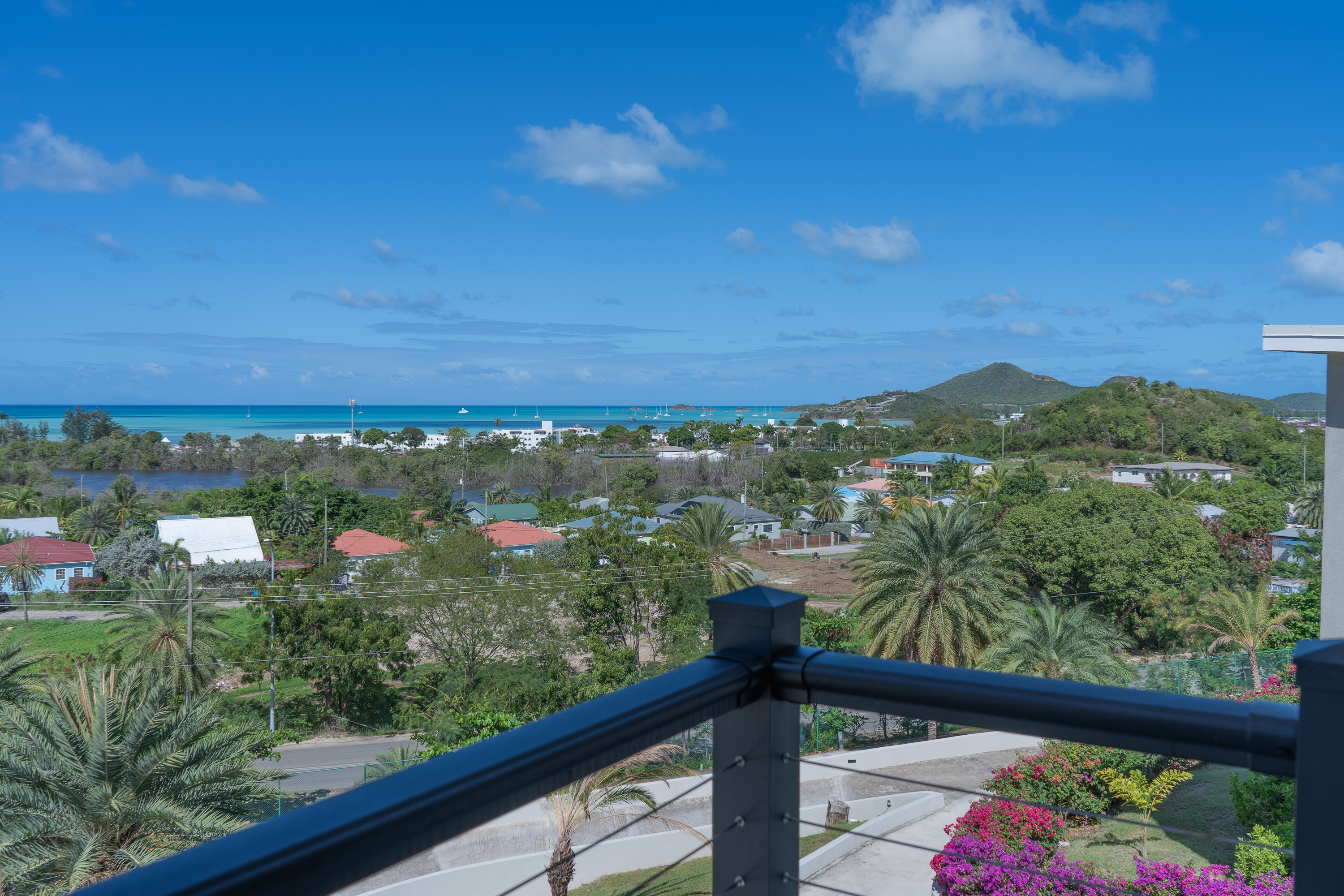 View from a balcony overlooking tropical greenery, scattered houses with red and white roofs, a calm blue sea with boats, and distant hills under a partly cloudy sky.