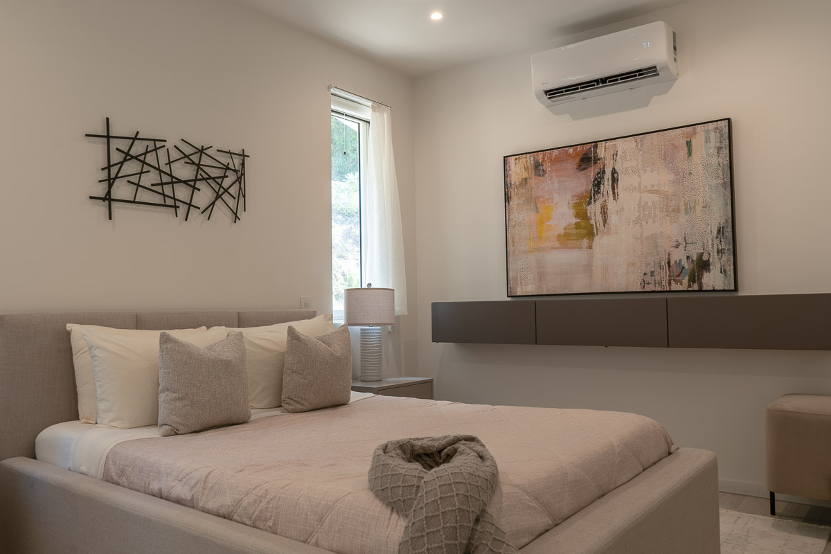 Modern bedroom with beige bed, neutral pillows, wall-mounted abstract painting, decorative wall art, lamp, window with white curtain, and air conditioner.