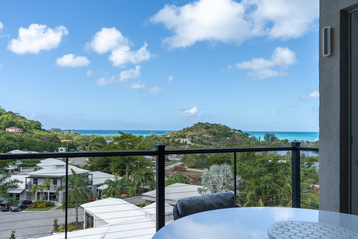 View from a balcony showing a round table with a cushioned chair, overlooking houses, greenery, hills, and a blue ocean under a partly cloudy sky.