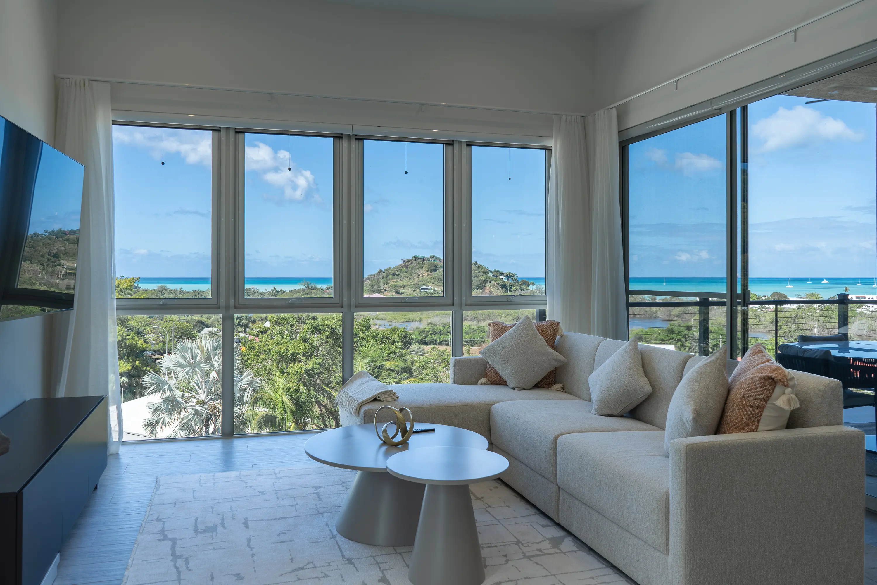 Modern living room with beige sectional sofa, two round coffee tables, and large windows showing ocean and greenery views.
