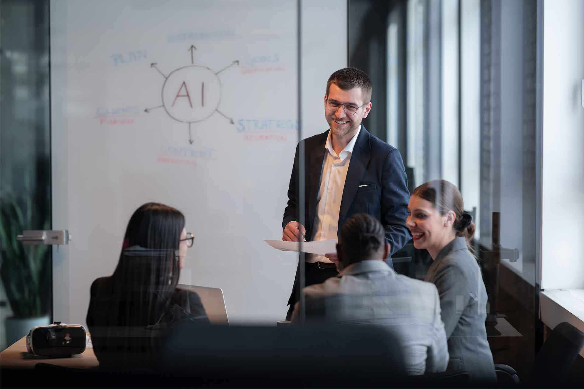 Four professionals have a meeting in a glass office, with "AI" and related terms written on a whiteboard behind them.