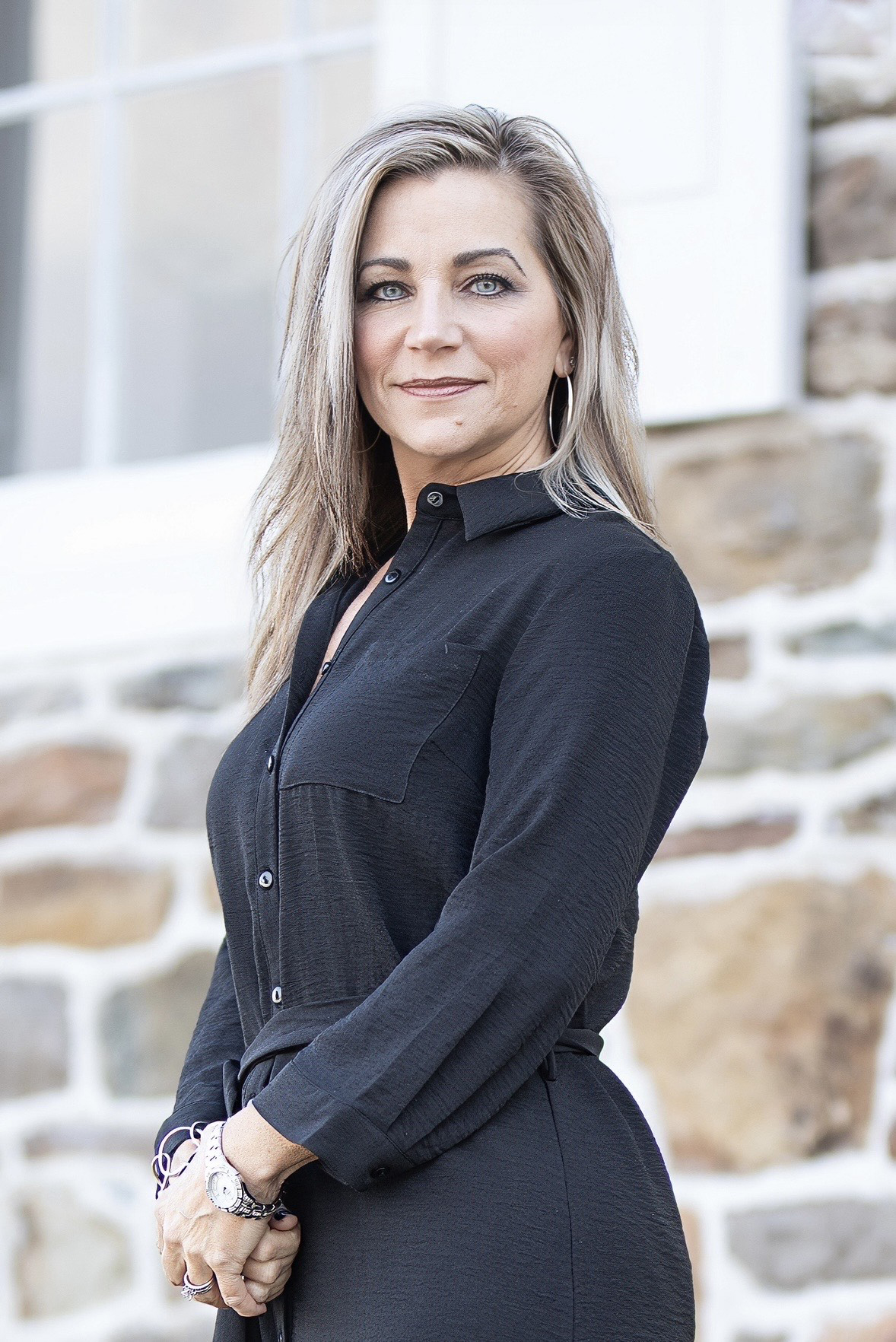 Portrait of a woman with long blonde hair wearing a black button-up shirt and hoop earrings, looking confidently at the camera.